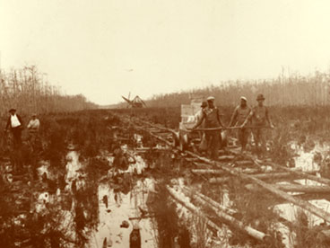Four workmen pulling loaded dynamite cart over a makeshift log tramway duing Tamiami Trail construction.  Dredge working in distance (center).  C. 1926