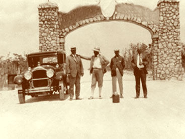 Four men posing by stone archway on Tamiami Trail which marked boundary between Collier and Dade County.  Automobile on left.  Pictured L to R:  Robert West, John Hachmeister, unidentified man and Attorney Patteau.  April 26, 1928.  (John Hachmeister Collection)